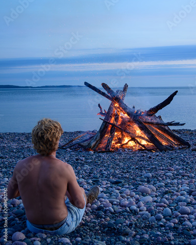 Person watches bonfire on Lake Superior beach vertical | A person sits before a glowing bonfire on a rocky beach along Lake Superior's north shore at dusk, in portrait orientation.