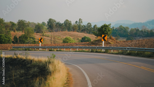 Curved rural highway with chevron warning signs and steel guardrail surrounded by dry countryside and trees. Scenic transportation infrastructure highlighting road safety, travel, navigation.