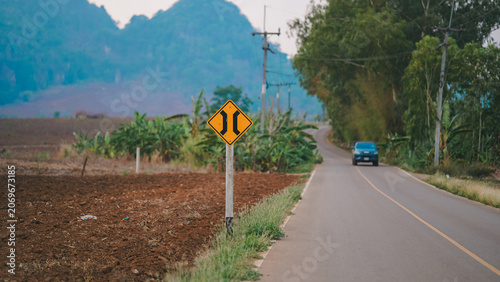 Road narrows warning sign beside a rural countryside road with passing vehicle, farmland and distant mountains. Scenic transportation landscape highlighting traffic safety, travel, infrastructure.
