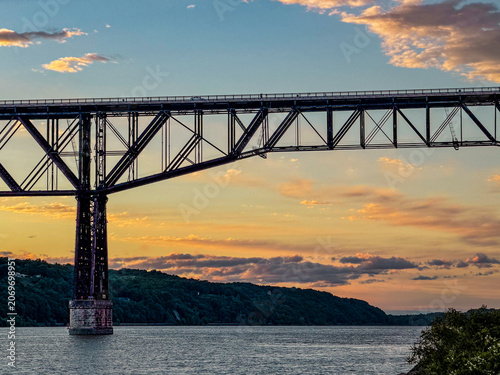Golden hour light bathes the Hudson River as the Walkway Over the Hudson stretches across calm waters, framed by rolling hills and scattered clouds, capturing a serene Poughkeepsie evening view