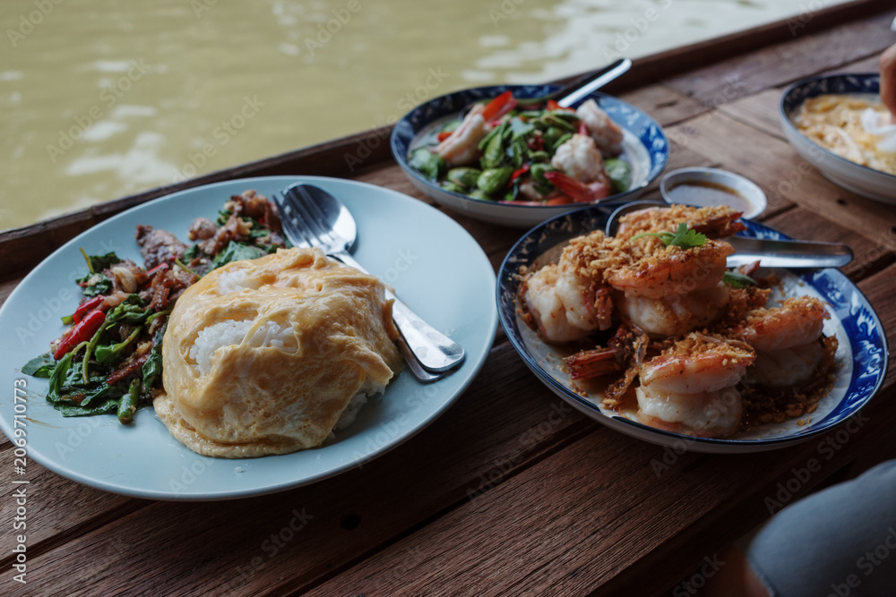 Fototapeta premium An eye-level photograph of a traditional Thai lunch set served on a wooden table beside a river, featuring a plate of rice topped with a creamy omelet alongside stir-fried shrimp dishes. 