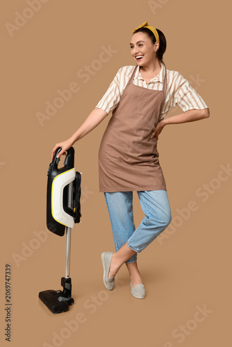Obraz na płótnie Portrait of happy young housewife in apron with vacuum cleaner on brown backgrou