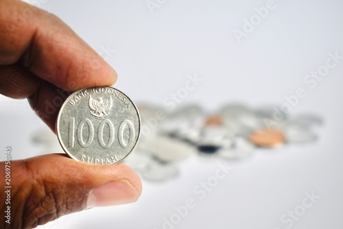 Fotografie Close-up of a hand holding a silver 1000 Indonesian Rupiah coin with a blurred background of scattered coins, representing payment and currency value