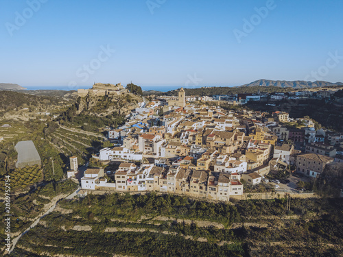 Aerial view of the historic town of Polop with traditional white houses, stone castle ruins, and terraced agricultural fields in Barony of Polop, Valencian Community, Spain.