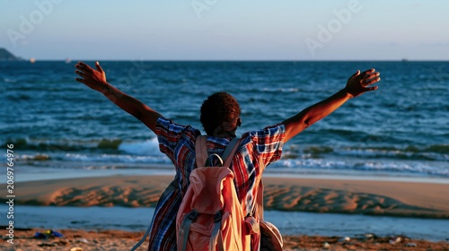 Young man with a backpack stands facing the ocean with arms wide open, enjoying the warm glowing sunset at the beach for travel and lifestyle content