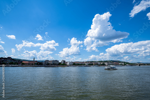 Wide view of the Danube River and Budapest waterfront in Hungary, with a passenger boat beneath a vivid blue sky filled with dramatic summer clouds.