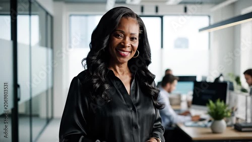 Smiling mature Black businesswoman standing in a modern corporate office with colleagues working in the background