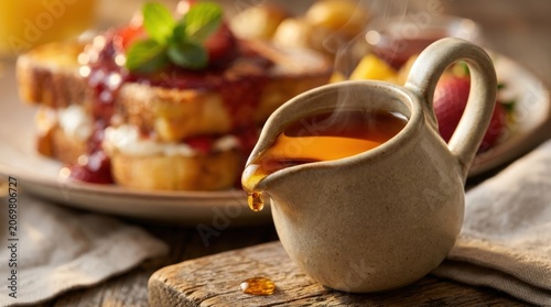 Steaming hot maple syrup being poured from a creamer onto a rustic wooden surface with french toast in the background