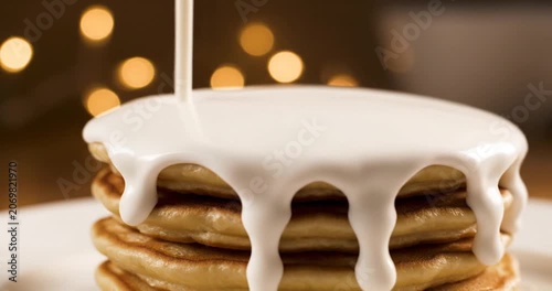 Stack of fluffy pancakes being drizzled with creamy topping against a blurred warm background