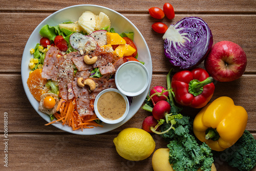 Fresh mixed vegetable and fruit salad bowl with crispy bacon, sesame dressing, and colorful ingredients on wooden background.