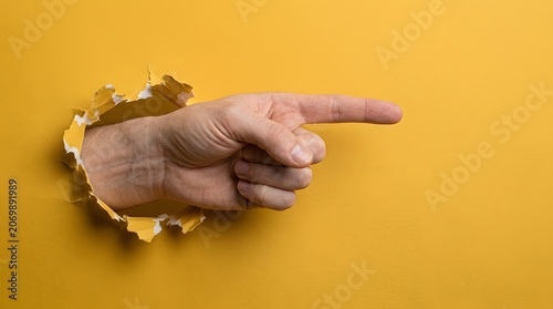 Pointing hand through torn yellow paper, human finger gesture breaking through background, directional sign concept for marketing and advertising