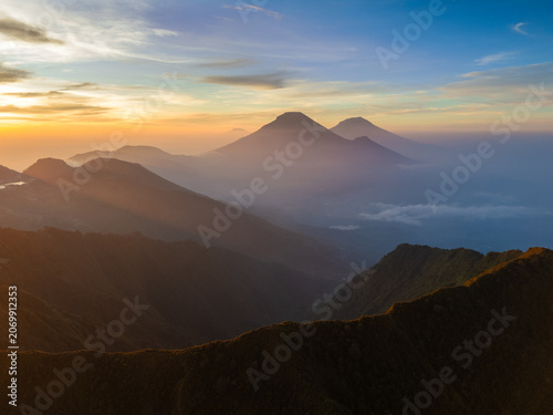 Aerial view of the mountainous landscape near Mount Bismo at sunrise, with golden light illuminating the misty peaks and deep valleys in Kecamatan Kejajar, Jawa Tengah, Indonesia.