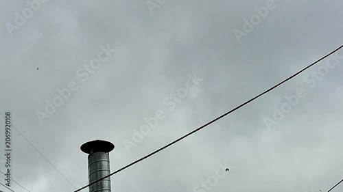 Storm clouds gather above an urban rooftop chimney and power lines, creating a dramatic city skyline scene. Moody weather, shifting light, and atmospheric skies capture the approach of a storm