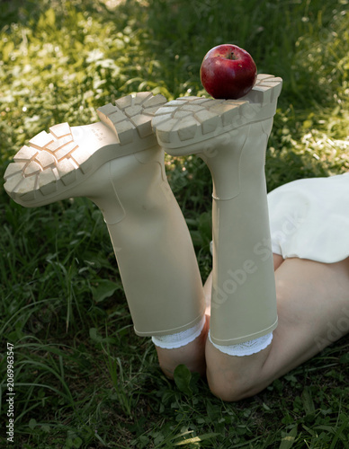 Creative portrait of woman in white lace outfit posing with red apples, balancing fruit on her head. Close up of girl with scraped, wounded knees covered with a green leaf in nature. Fairytale concept