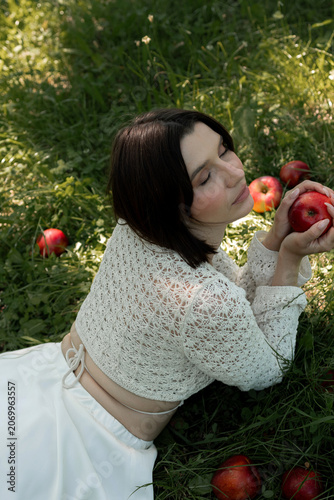 Creative portrait of woman in white lace outfit posing with red apples, balancing fruit on her head. Close up of girl with scraped, wounded knees covered with a green leaf in nature. Fairytale concept