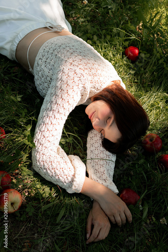 Creative portrait of woman in white lace outfit posing with red apples, balancing fruit on her head. Close up of girl with scraped, wounded knees covered with a green leaf in nature. Fairytale concept
