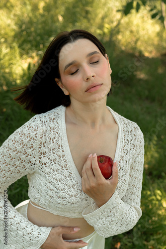 Creative portrait of woman in white lace outfit posing with red apples, balancing fruit on her head. Close up of girl with scraped, wounded knees covered with a green leaf in nature. Fairytale concept