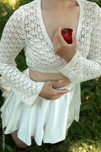 Creative portrait of woman in white lace outfit posing with red apples, balancing fruit on her head. Close up of girl with scraped, wounded knees covered with a green leaf in nature. Fairytale concept