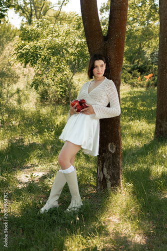 Creative portrait of woman in white lace outfit posing with red apples, balancing fruit on her head. Close up of girl with scraped, wounded knees covered with a green leaf in nature. Fairytale concept