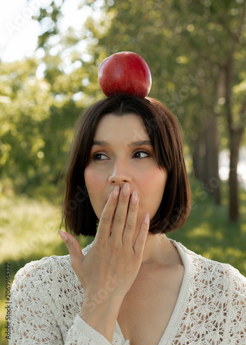Creative portrait of woman in white lace outfit posing with red apples, balancing fruit on her head. Close up of girl with scraped, wounded knees covered with a green leaf in nature. Fairytale concept