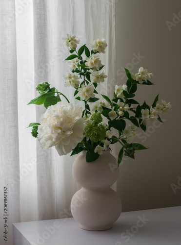 Bouquet of jasmine and white peonies in a ceramic vase on a white wooden table in the living room