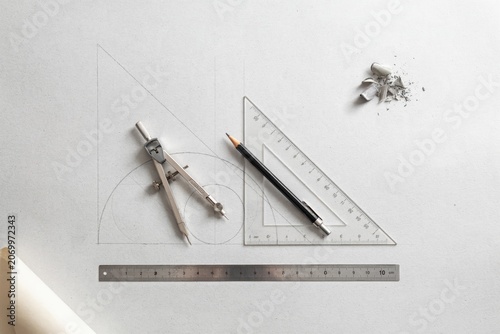 Technical setup on a white surface featuring a metal compass, a transparent plastic triangle ruler, a pencil, and a metal ruler