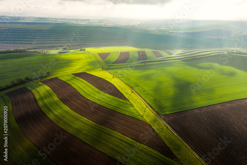 Aerial view of rolling agricultural fields with alternating strips of green crops and dark soil under dramatic sunlight and cloud shadows, Klobouky u Brna, Jihomoravsky kraj, Czechia.