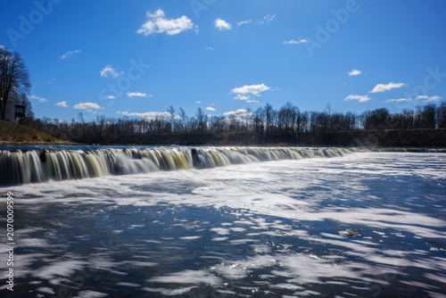 View of Ventas Rumba, the widest waterfall in Europe, with flowing water cascading over the limestone ledge under a clear blue sky in Kuldiga, Kuldigas novads, Latvia.