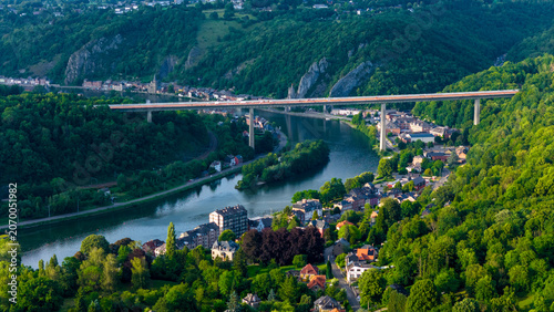 Aerial view of the Charlemagne Bridge crossing the Meuse River, surrounded by the historic town and lush green forests on the valley slopes in Dinant, Wallonia, Belgium.