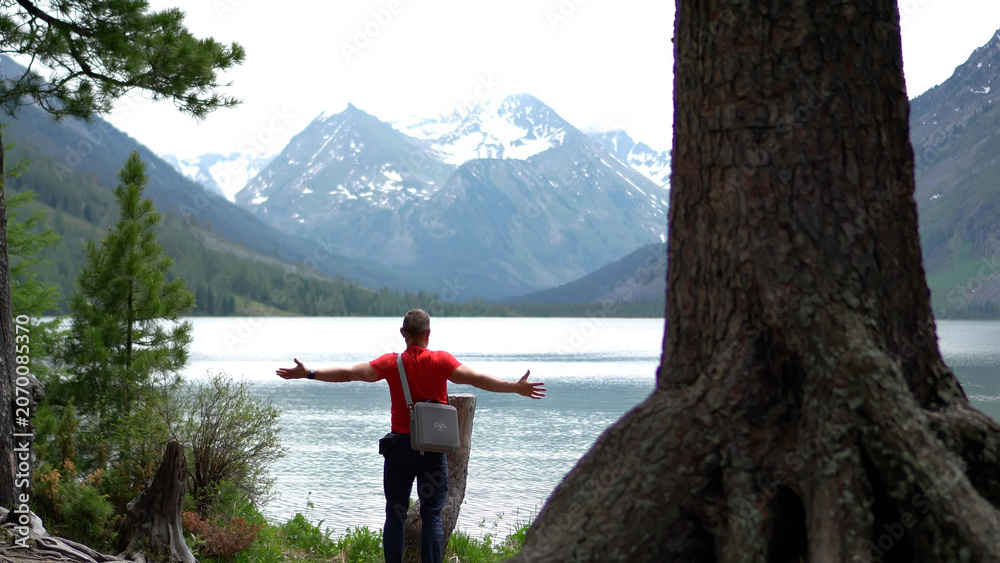 Obraz premium A male adventurer in a red t-shirt standing with outspread arms on an alpine lake shore, celebrating freedom against a majestic snow-capped mountain backdrop.
