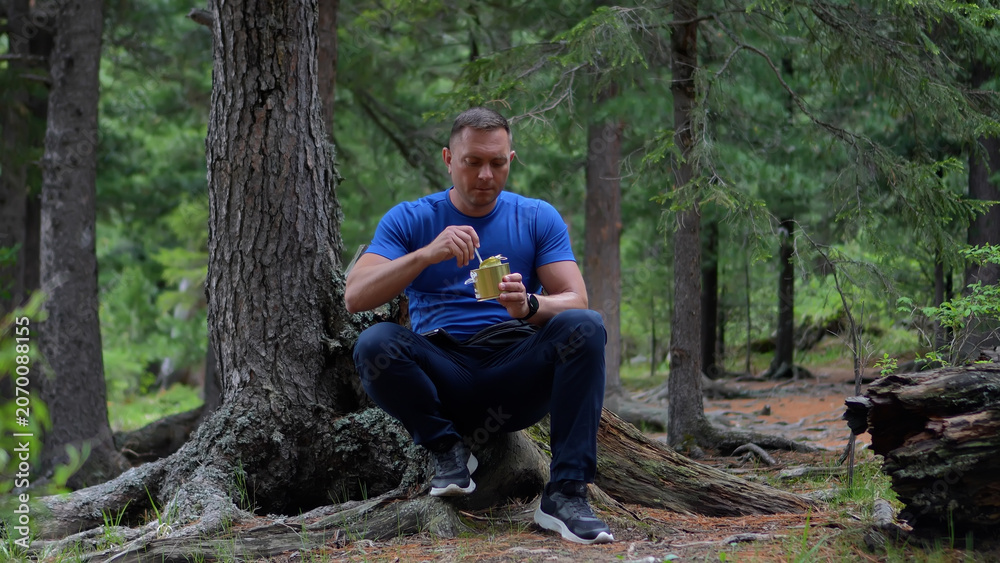 Fototapeta premium A focused male hiker in a blue t-shirt sitting on cedar roots and opening a tin can of food in the Altai forest.