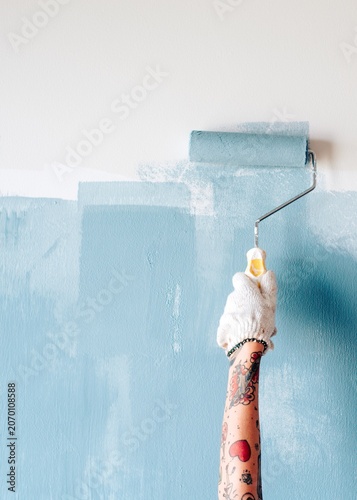 Принти на полотні Tattooed arm painting a wall blue with a roller, wearing a glove.