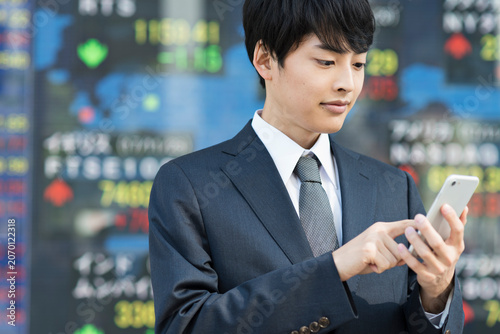 Cuadro en lienzo 株価ボード前でスマートフォンを操作する20代男性ビジネスマン