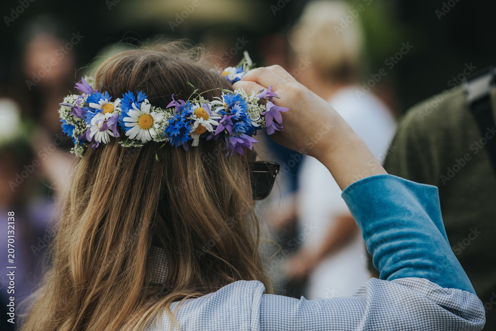 Naklejka premium Young woman with long brown hair wearing a floral crown made of daisies and wildflowers adjusts her hair while standing outdoors in a vibrant festival setting
