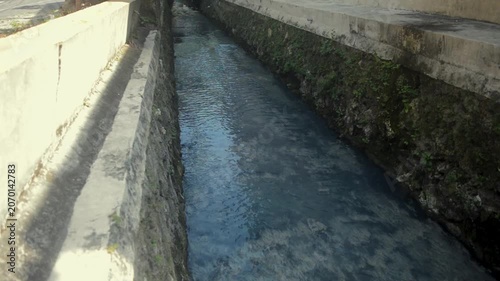 Narrow Concrete Water Canal With Flowing Dark Water And Green Mossy Sides In A Sunny Outdoor Setting