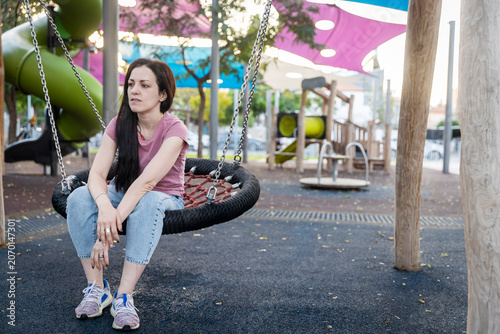 Canvastavla A woman with long dark hair, a pink t-shirt, light blue jeans, and multi-colored sneakers, sits thoughtfully on a large round net swing at an outdoor playground