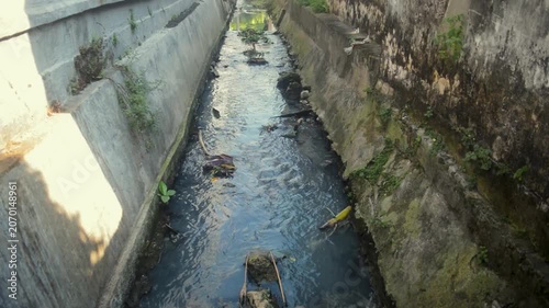 Narrow Urban Drainage Channel With Murky Flowing Water And Weathered Concrete Walls In City Alley