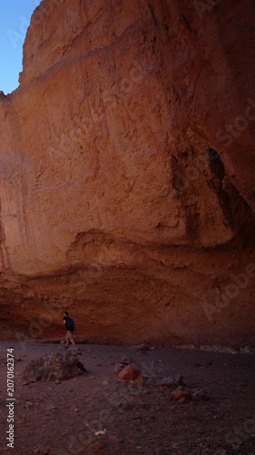 Vertical Screen: Solo hiker with backpack explores massive weathered red sandstone cliff walls in arid desert canyon during golden hour light with clear blue sky and scattered rocks on dusty ground.