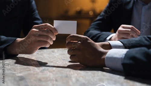 Diverse professional business team members in elegant suits collaborating and holding out a blank white business card for mockup placement in a bright modern office setting with copy space.