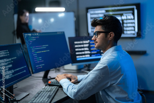 Tableau sur toile Young South Asian man in glasses works at a multi-monitor setup with code and data screens as a female colleague stands in the background