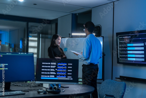South Asian man presents at a whiteboard beside a wall screen showing a data dashboard while a woman listens in a dark blue office meeting room.