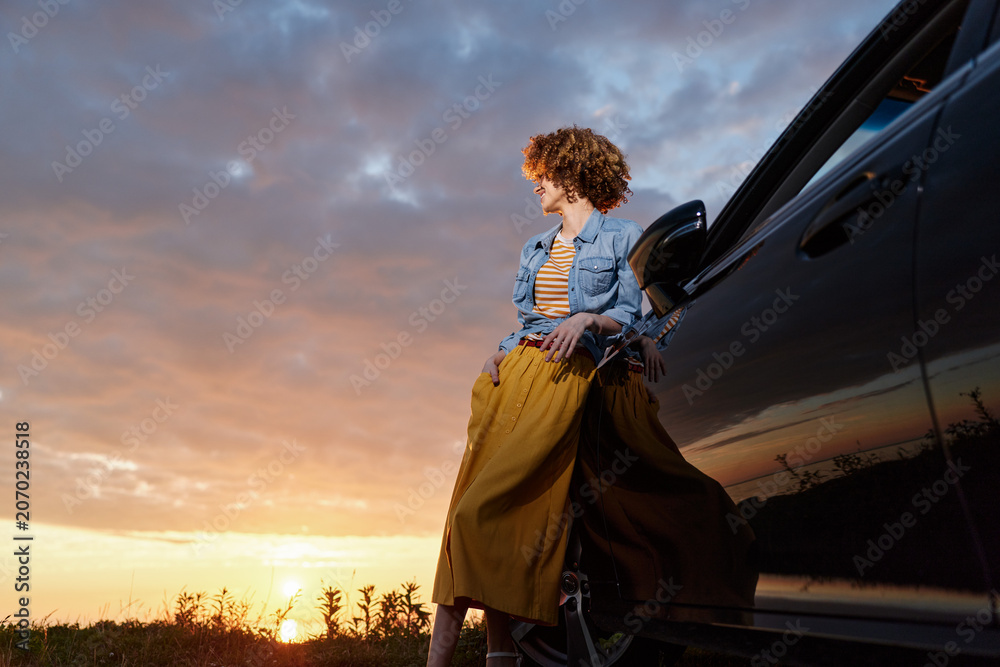 Fototapeta premium Young woman with curly hair sits beside a car at sunset, wearing a denim jacket and yellow skirt, relaxed and thoughtful, with warm light curling over the horizon.