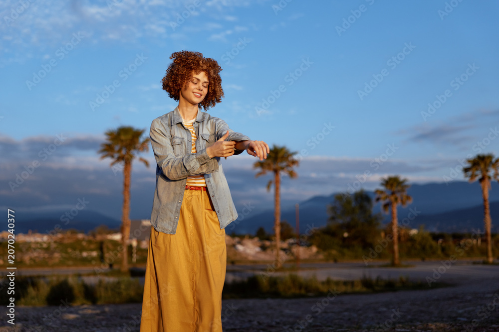 Fototapeta premium Cheerful woman outdoors during golden hour, wearing denim jacket and yellow dress, smiling with relaxed posture, warm light, palm trees and distant mountains in background