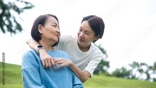 Photography A smiling Asian daughter lovingly embraces her senior mother from behind in a green park