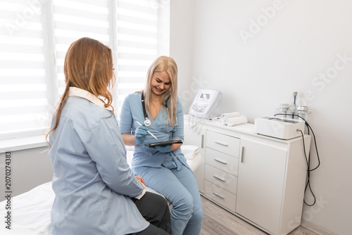 Young woman doctor in blue uniform attentively listens to female patient, reviewing medical records on tablet. Contemporary clinic interior emphasizes trust, care, and advanced healthcare services
