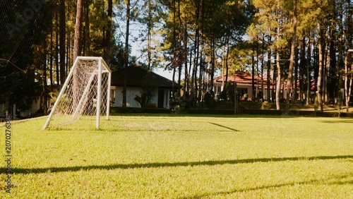A white soccer goal stands on a green field in a forest clearing with trees and a house in background