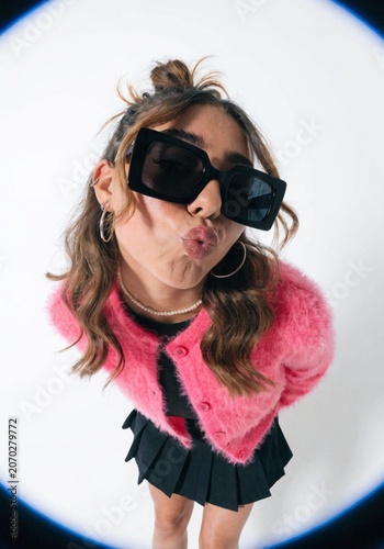 A stylish young woman poses playfully while pouting her lips, wearing a fluffy pink cardigan and dark sunglasses captured through a distorted fisheye lens perspective against a plain white background