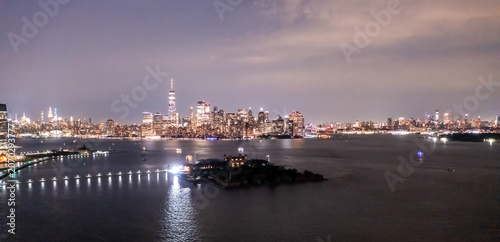 New York, New York, United States - 11 August 2021: Aerial view of the Manhattan skyline at night with Ellis Island in the foreground and the Hudson River reflecting city lights.