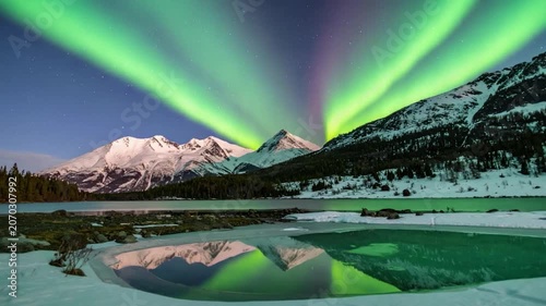 Green aurora borealis lights over snowy mountains reflecting in a lake