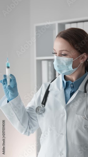 Confident woman doctor wearing protective mask and gloves examines syringe with vaccine in bright hospital office. Medical professional ready for vaccination or clinical treatment procedure